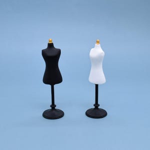 May include: Two miniature dress forms, one black and one white, stand side-by-side against a light blue background. Each form has a gold-colored top and a black stand. The forms are designed to resemble a classic dressmaker's mannequin.