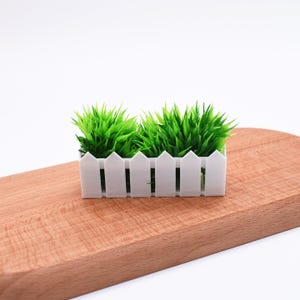 Può includere: Un piccolo vaso bianco a forma di staccionata, riempito di erba artificiale verde brillante. Il vaso rettangolare è posto su un tagliere di legno, creando un contrasto di trame e colori. L'erba verde brillante aggiunge un tocco naturale.