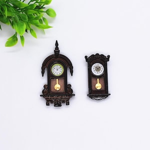 Puede incluir: Dos relojes de pared en miniatura de estilo antiguo. Los relojes son marrones con péndulos y esferas doradas. Las esferas tienen números y manecillas negras. Un reloj tiene una esfera verde y el otro una blanca. Los relojes están sobre un fondo blanco.