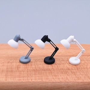 May include: Three miniature desk lamps in grey, black, and white. Each lamp has a white lampshade and adjustable arm. The lamps are on a wooden surface, with a light grey background.