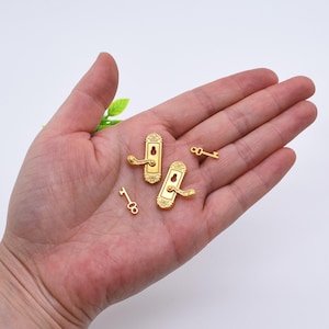 May include: Miniature gold-colored door handles and keys. The door handles have a detailed design with a curved handle and a keyhole. Two small keys are also visible. The items are likely for a dollhouse or craft project.