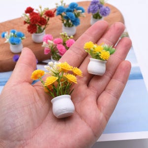 Może przedstawiać: Miniaturowe kwiaty doniczkowe w różnych kolorach, w tym czerwonym, niebieskim, żółtym, różowym i fioletowym. Małe, sztuczne rośliny znajdują się w białych ceramicznych doniczkach. Kwiaty są ułożone na drewnianej powierzchni.