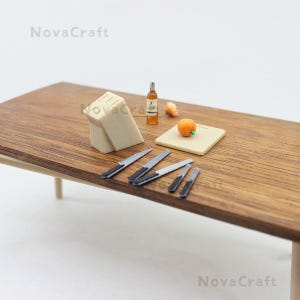 Op de afbeelding: Miniatuurkeuken met een houten tafel, een messenblok en verschillende kleine messen. Een fles, een snijplank met een sinaasappel en een stuk voedsel staan ook op de tafel. De scène is een miniatuurweergave van een keuken.