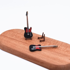 以下が含まれることがあります： 木製の表面に置かれた、黒と赤のミニチュアのエレキギター。1つのギターは小さな木製のスタンドに立っており、もう1つは平らに置かれています。小さな木製のギタースタンドも存在します。