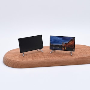 May include: Two miniature televisions, one with a blank black screen and the other showing a scenic image of a bridge and city lights. Both are mounted on silver stands and set on a wooden oval base.