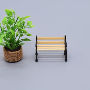 May include: Miniature park bench with a light brown seat and black frame. The bench has horizontal slats for the seat and back. A small potted plant with green leaves sits to the left of the bench.