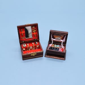 Puede incluir: Dos cajas de madera en miniatura, una abierta con espejo, tijeras y joyas sobre un forro de terciopelo rojo. La otra caja contiene carretes de hilo y artículos de costura. Ambas cajas son marrones con herrajes dorados.