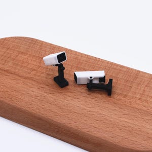 以下が含まれることがあります： 2つのミニチュア防犯カメラ。1台のカメラは黒いスタンドに取り付けられ、もう1台は黒いブラケットに取り付けられています。どちらのカメラも白で、黒いレンズが付いています。カメラは木製の表面にあります。