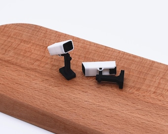 Könnte beinhalten: Zwei Miniatur-Überwachungskameras mit weißen rechteckigen Gehäusen und schwarzen Montagehalterungen. Eine Kamera steht aufrecht auf ihrem Sockel, während die andere horizontal liegt und den schwarzen Stützarm sowie die Basisstruktur zeigt.
