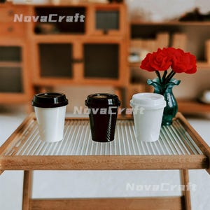 Peut inclure: Trois petites tasses à café miniatures, dont deux blanches et une marron foncé, avec des couvercles noirs et blancs. Elles sont posées sur une petite table en bois. Un vase avec des fleurs rouges est en arrière-plan. Le texte "NovaCraft" est visible.