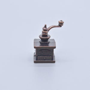 May include: A small, bronze-coloured coffee grinder with a hand crank. The grinder has a square base with a small drawer and the word "CAFE" is visible on the front. The crank arm extends to the right, and the design is detailed.
