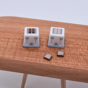Op de afbeelding: Twee miniatuur witte broodroosters met grijze afwerking en bruine toast worden gepresenteerd op een houten snijplank. De broodroosters hebben sleuven voor de toast en kleine details. De scène speelt zich af op een lichtgekleurd houten oppervlak.