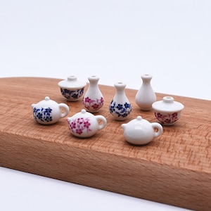 Puede incluir: Colección de miniaturas de cerámica: teteras, jarrones y cuencos con tapa. Las piezas de porcelana blanca están decoradas con motivos florales azules y rosas. Dispuestas sobre una superficie de madera.