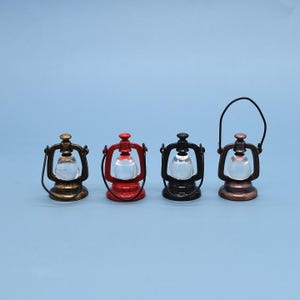 May include: Four miniature lanterns in varying colors: gold, red, black, and bronze. Each lantern has a clear glass globe and a metal frame with a handle. The lanterns are arranged on a light blue background.