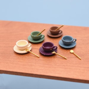 Può includere: Servizio da tè in miniatura con sei tazzine, piattini e cucchiaini colorati. Le tazzine sono di vari colori, tra cui crema, verde, marrone, blu e viola. I cucchiaini sono dorati. Il set è disposto su una superficie di legno.