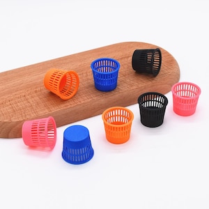 May include: A collection of miniature, colorful trash cans in orange, blue, pink, and black. The small, cylindrical containers are arranged on a wooden cutting board. The cans have a slotted design.