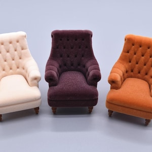 May include: Three miniature armchairs in cream, burgundy, and orange. Each chair has a tufted back and armrests, with small wooden legs. The chairs are arranged side by side against a light grey background, showcasing their plush, velvet-like texture.