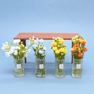 Op de afbeelding: Vier kleine glazen vazen met verticale ribbels, elk met kunstbloemen in verschillende kleuren: wit, lichtblauw, geel en oranje. Op de achtergrond een kleine houten tafel, tegen een lichtblauwe achtergrond.