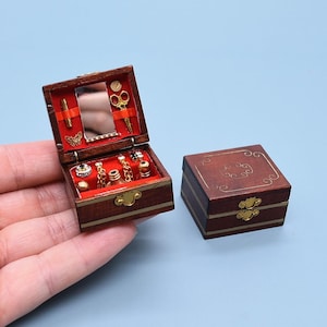 Op de afbeelding: Miniatuur houten sieradendoosje met een rode fluwelen binnenkant, gevuld met kleine goudkleurige accessoires. De open doos toont een spiegel, schaar en andere kleine items. Een tweede, gesloten doos is ook aanwezig.