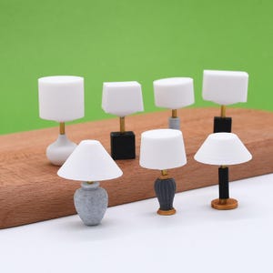 May include: A collection of miniature lamps with white shades and various base designs. The bases include gray, black, and gold accents. The lamps are arranged on a wooden surface against a green backdrop, showcasing a variety of styles.