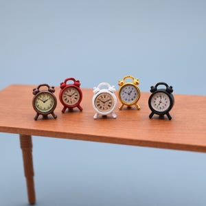 Könnte beinhalten: Fünf Miniatur-Wecker in verschiedenen Farben: Braun, Rot, Weiß, Gold und Schwarz. Jede Uhr hat ein rundes Zifferblatt mit schwarzen Ziffern und Zeigern. Die Uhren werden auf einer Holzoberfläche vor hellblauem Hintergrund ausgestellt.