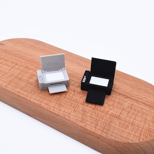 Può includere: Due stampanti in miniatura, una grigia e una nera, sono esposte su una superficie di legno. Ogni stampante ha la parte superiore aperta e un vassoio esteso. La stampante grigia ha un piccolo display laterale. Le stampanti sono modelli piccoli e dettagliati.