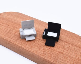 May include: Two miniature printers, one gray and one black, are displayed on a wooden surface. Each printer has an open top and a tray extended. The gray printer has a small display on the side. The printers are small, detailed models.