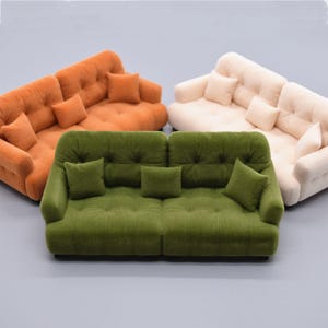 Puede incluir: Sofás en miniatura en colores naranja, verde y crema. Cada sofá tiene un diseño capitoné y viene con cojines cuadrados a juego. Los sofás están dispuestos sobre una superficie gris, mostrando una colección de muebles de casa de muñecas.