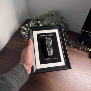 May include: Framed artwork featuring a car key. The key is silver with black accents and the Porsche logo. The frame has a black border and a white mat. Text at the top reads "NO RISK, NO STORY."