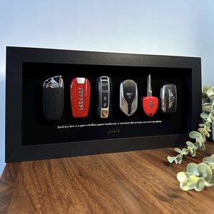 Può includere: Un'esposizione incorniciata in nero con sei chiavi di auto, tra cui Lamborghini, Ferrari e Porsche. Le chiavi sono disposte su uno sfondo nero. Il testo in basso recita: "Ogni chiave qui è una silenziosa ribellione contro la mediocrità, un'affermazione che la media non è mai stata un'opzione."