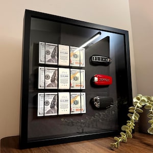 Peut inclure: Un présentoir encadré noir avec trois piles de billets de 10 000 $ US, maintenues par des bandes noires. Le présentoir contient également trois clés de voiture : noire, rouge et argentée. Le fond est noir.