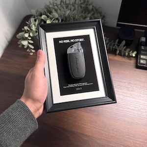 May include: Framed artwork featuring a Lamborghini car key on a black background. The key is dark gray with silver accents, and the frame is black. The text "NO RISK, NO STORY" is at the top. The artwork is displayed on a wooden surface.
