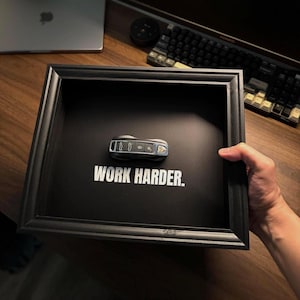 Może przedstawiać: Czarna oprawiona ekspozycja z kluczykiem do samochodu i napisem "WORK HARDER". Kluczyk jest srebrno-czarny. Ekspozycja znajduje się na drewnianej powierzchni.