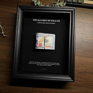 Peut inclure: Une œuvre d'art encadrée en noir avec un billet de 100 dollars plié. Le texte "THE ILLUSION OF WEALTH" est visible. Le fond est noir. Le cadre est posé sur une surface sombre.