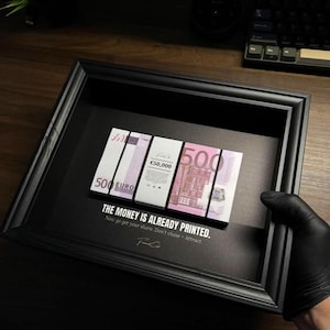 Può includere: Un'opera d'arte incorniciata in nero con una pila di banconote da 500 euro, tenute insieme da un elastico nero. L'opera include il testo "THE MONEY IS ALREADY PRINTED." e una firma.