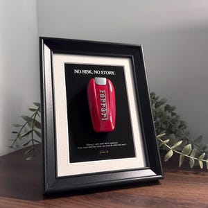 May include: Framed artwork featuring a red Ferrari key on a black background, with the text "NO RISK, NO STORY." and a quote. The key is centered, and the frame is black with a white mat.
