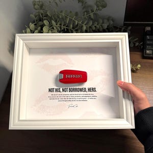 May include: A framed artwork featuring a red Ferrari key. The key is centered on a white background with the text "NOT HIS, NOT BORROWED, HERS." and a lipstick kiss mark. The frame is white.