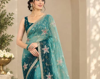 May include: A teal and light turquoise saree featuring a gradient color transition, adorned with embroidered star-shaped floral motifs in white and pink, accented with silver sequins and a decorative metallic border trim.