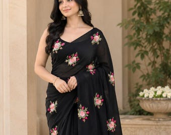 May include: A black saree featuring a sleeveless blouse and a long, flowing drape. The fabric is adorned with scattered, embroidered floral motifs in shades of pink, green, and white. The design includes a V-neckline and a sheer, lightweight texture.