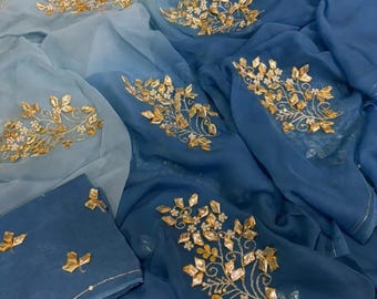 May include: A flowing, layered fabric in shades of blue, from light to deep indigo. The fabric is adorned with gold-colored floral embroidery. The design features intricate leaf and petal patterns, creating a luxurious and elegant aesthetic.