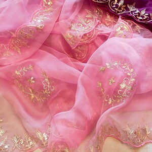 May include: A draped pink fabric with gold floral embroidery. The delicate material is adorned with intricate gold detailing along the edges and across the surface. A section of deep purple fabric with gold embroidery is also visible.