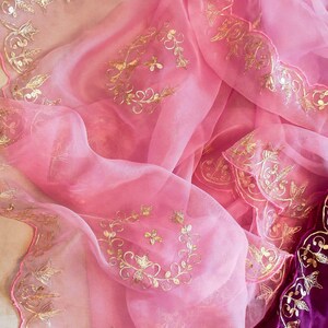 K&ouml;nnte beinhalten: Ein durchscheinender, rosa Stoff mit goldfarbenen, floralen Stickereien. Der Stoff hat einen gewellten Rand und ist drapiert, wodurch ein tiefvioletter Samtstoff sichtbar wird. Die goldene Stickerei verleiht dem zarten rosa Material Eleganz.