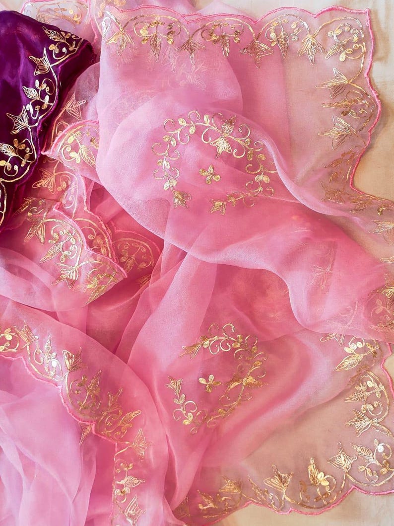 K&ouml;nnte beinhalten: Zartrosa, durchsichtiger Stoff mit goldfarbenen Blumenstickereien. Der Stoff ist drapiert und zeigt einen tiefvioletten Stoff mit goldfarbenen Stickereien. Die Kanten sind gewellt. Das Design ist filigran und verziert, mit Fokus auf den goldenen Details.