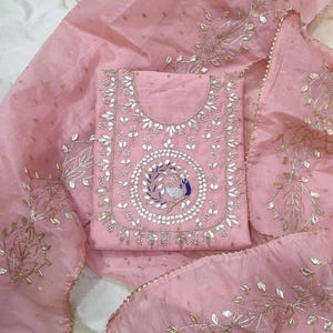 Pode incluir: Um terno salwar kameez indiano rosa claro com bordados intrincados. O terno apresenta um design de pavão detalhado na frente, cercado por padrões florais e de folhas. O dupatta e o terno são feitos de um tecido transparente com detalhes dourados.