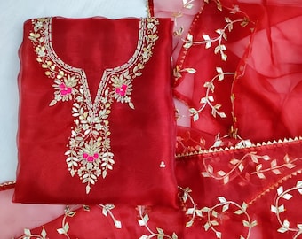 May include: A bright red salwar kameez outfit. The top has detailed gold and pink floral embroidery around the neckline and centre. The sheer dupatta and top are decorated with gold leaf patterns. A traditional Indian outfit.