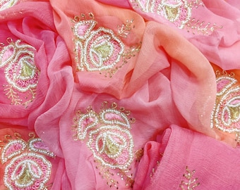 May include: A close-up of a draped fabric in shades of pink and peach. The fabric is adorned with embroidered floral designs in pink, gold, and white. The embroidery features intricate details and a beaded outline, creating a luxurious look.