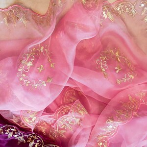 K&ouml;nnte beinhalten: Ein flie&szlig;ender, durchsichtiger rosa Stoff mit aufwendiger goldener Stickerei. Der Stoff ist drapiert und gefaltet und zeigt die zarten Blumen- und Blattmuster. Ein tiefvioletter Stoff mit goldener Stickerei ist unten sichtbar.