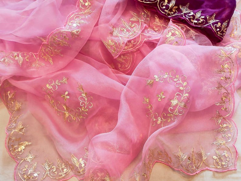 K&ouml;nnte beinhalten: Ein flie&szlig;ender rosa Stoff mit goldenen Blumenstickereien. Das zarte Material ist drapiert und zeigt die komplizierten goldenen Details an den R&auml;ndern und &uuml;ber die Oberfl&auml;che verteilt. Ein Abschnitt aus tiefviolettem Stoff mit Goldstickerei ist sichtbar.