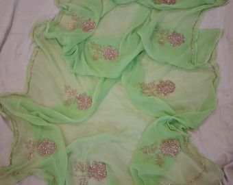 May include: A light green sheer fabric with embroidered floral designs and a gold trim. The fabric is draped and folded, showcasing the delicate embroidery and the flowing nature of the material. The floral designs are clustered in various areas.