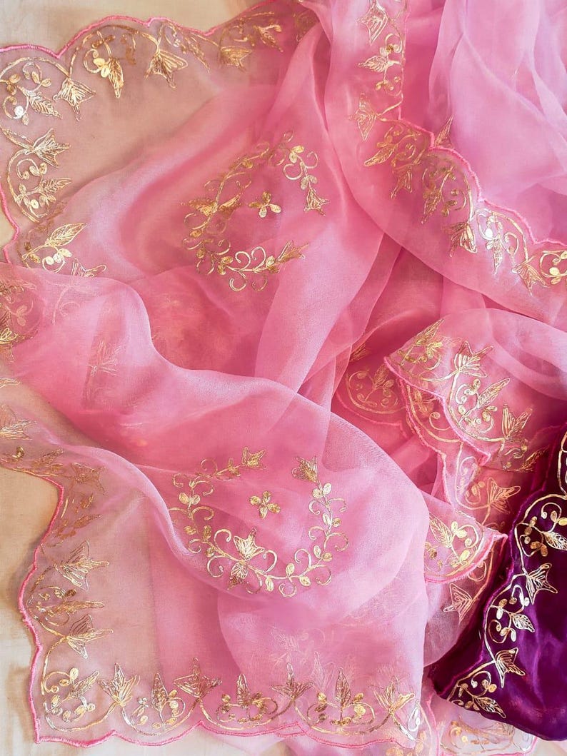 K&ouml;nnte beinhalten: Ein durchscheinender rosa Stoff mit goldener Stickerei. Der Stoff ist drapiert und zeigt aufwendige Blumen- und Blattmuster. Die Kanten sind gewellt, und ein tiefvioletter Samtstoff ist sichtbar.